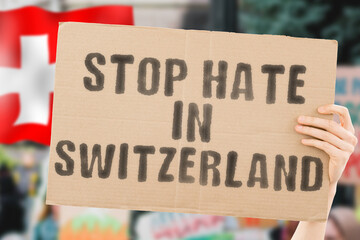 The phrase " Stop hate in Switzerland " on a banner in men's hand with blurred Swiss flag on the background. Gay. Human rights. Equality. Social issues. Society. Problem. Conflict