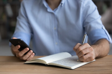 Crop close up of young Caucasian man handwrite in notebook using modern cellphone gadget application online. Male write in notepad make list or plan with smartphone, study distant on device.