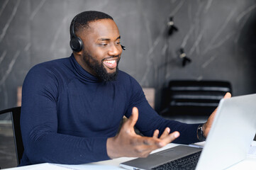 Focused young businessman in eyewear wearing headphones, holding video call with clients on laptop. Concentrated african man in glasses giving online educational class lecture, consulting customer