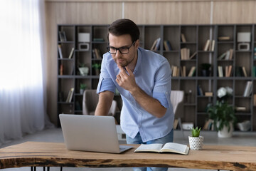 Pensive Caucasian man work online on laptop from home office make notes consult client or customer. Thoughtful young male look at computer screen make decision or plan, study distant on gadget.