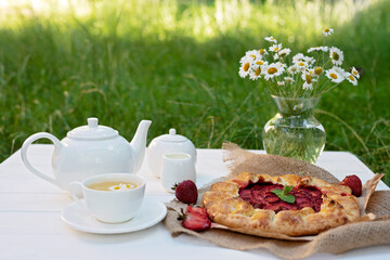 Freshly baked homemade galette or open strawberry pie, a cup of herbal tea, a teapot and a vase with a bouquet of daisy flowers, summer outdoor food. Soft selective focus.