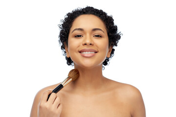 beauty, cosmetics and people concept - portrait of happy smiling young african american woman with make up brush applying shimmer to her skin over white background