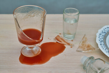 A broken glass, red tomato juice spilled on the table next to a bottle of vodka