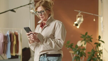 Elegant senior woman standing in photo studio and typing on smartphone while working in fashion agency