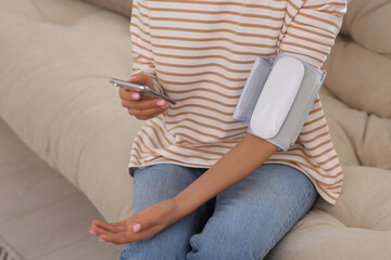 Woman checking blood pressure with modern monitor and smartphone indoors, closeup