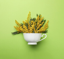 Beautiful floral composition with mimosa flowers and cup on green background, top view