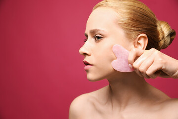 Obraz na płótnie Canvas Beautiful young woman doing facial massage with gua sha tool on pink background, closeup. Space for text