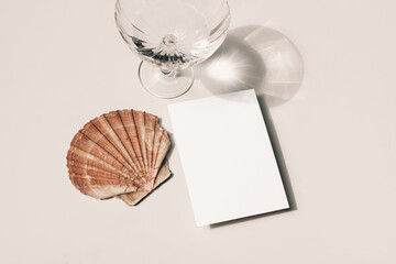 Modern summer still life. Glass of water, cocktail with long shadows. Oyster sea shell on beige table background in sunlight. Blank greeting card mockup. Flat lay, top view. Birthday concept.