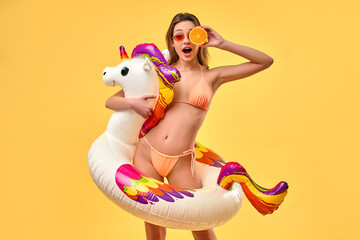 Naklejka premium Cute woman in swimsuit with unicorn inflatable multicolored circle and orange slice isolated on yellow background. Summer time. Rest, vacations, travel.