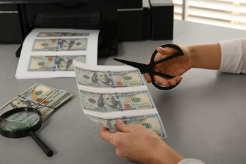 Counterfeiter cutting dollar banknotes with scissors at table indoors, closeup. Fake money concept