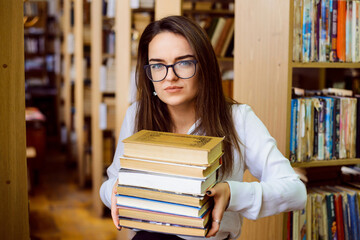 Female student with a big pile of books in the library. Bewildered girl preparing for examinations, sick and tired of constant hard learning