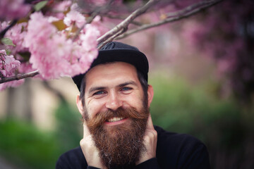 A cheerful bearded man with a long mustache and gray hair in a dark sweater and baseball cap laughs near the cherry blossoms in spring.