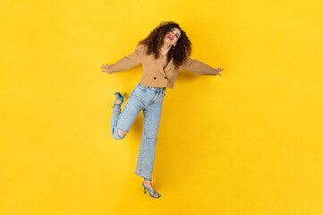 Curly girl in trendy outfit of jeans and jacket posing on the yellow background in the studio. Happy woman facial expression. Outfit of the day.