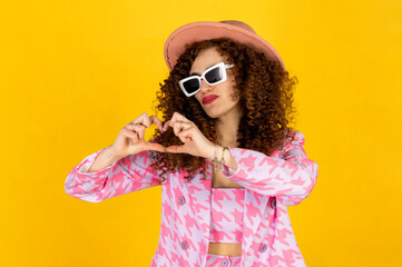 Funny and cool girl with curly hair in a pink outfit with hat posing. Isolated on yellow background. Showing heart sign with hands.