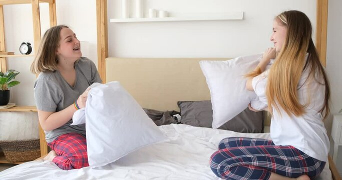 Two joyful young women fight pillows in the bedroom, slow motion