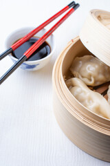 Top view of open steamer with gyozas, soy sauce in bowl with chopsticks, on white wood, vertical, with copy space