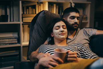spa weekend. couple with applied cosmetic face mask on their faces, relaxing sofa at their home. evening scene