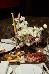 table in the restaurant. there is a bouquet of white flowers on the table, crispy long bread sticks in glasses and a plate of cold cuts and cheeses