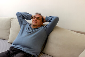 Retired man sitting on sofa, thinking about the future. Mindful old man daydream on cozy sofa in modern living room. Happy senior retired 70s grey haired man relaxing and enjoying peaceful lazy moment