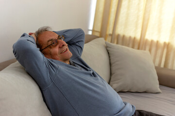 Obraz premium Peaceful older man feel relax on comfortable sofa in living room, calm mature male rest on cozy sofa, enjoy weekend at home. Side view of a 70s man with hands behind head enjoy at home, leaned on sofa
