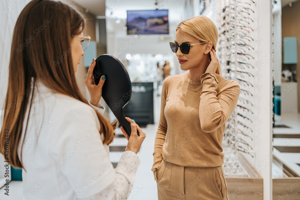 Poster beautiful and fashionable woman choosing eyeglasses frame in modern optical store. female seller spe