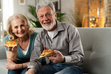 Cheerful husband and wife sitting on sofa at home. Happy senior woman and man eating pizza while...