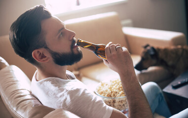 Handsome young smiling man is watching films on TV and drinks beer with snack while resting on the couch