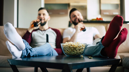 Two young happy bearded friends watching TV or some sport match while sitting on the couch at home on weekend and drinking beers and eating snacks