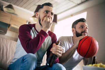 Two excited funny young friends fans of basketball watching TV match and shouting while resting on the couch