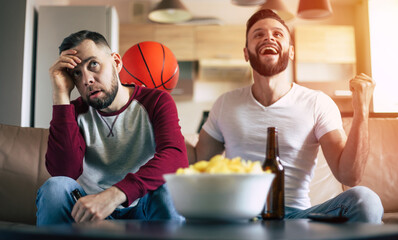 Two excited funny young friends fans of basketball watching TV match and shouting while resting on the couch