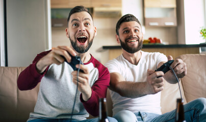 Excited smiling men playing in video games on tv at home on the couch. Friends with joysticks play game with happy emotions on faces © My Ocean studio