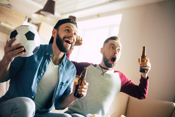 Funny excited hipster bearded friends with snacks and beers watching football on tv while sitting...