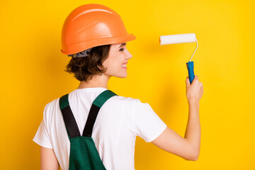 Back view photo of happy cheerful smiling woman builder in helmet painting walls isolated on yellow...