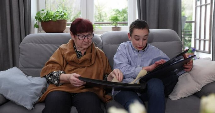 Senior old woman sitting with grandson at grey sofa at home and watching old photos in the big photo album emotionally.
