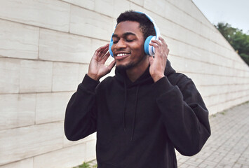 Portrait of smiling young african man in wireless headphones enjoying listening to music wearing a black hoodie on a city street