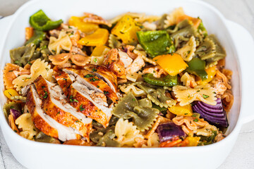 Farfalle pasta with baked vegetables and chicken in white dish, gray background.