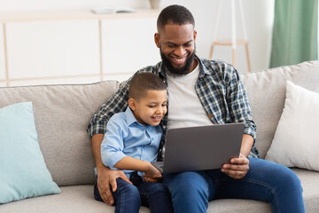 Fototapeta premium African Daddy Embracing Son Sitting With Laptop Watching Cartoons Indoor