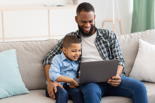 African Daddy Embracing Son Sitting With Laptop Watching Cartoons Indoor