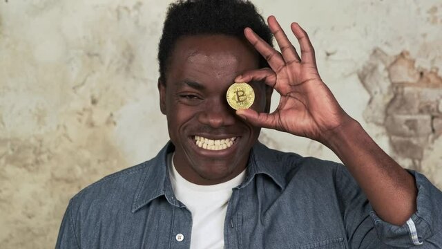 Funny african american man with charming smile standing covering one eye with golden bitcoin close-up. Cryptocurrency, blockchain, defi concept