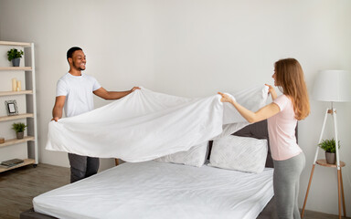 Fototapeta premium Cheerful multiracial couple making bed in morning, having fun at home
