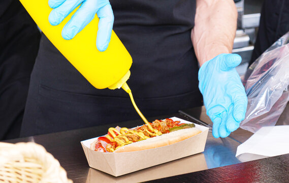 Street food cart worker prepares a hot dog. Gloved hands add mustard to the hot dog. Traditional takeaway, street food, snacks.
