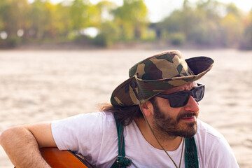 Hippy man with hat beard and glasses playing guitar outdoors on the beach