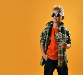 Calm rich teenager boy, lottery winner in round sunglasses, checkered plaid shirt and jeans stands holding fan of dollars cash isolated over yellow background with copy space