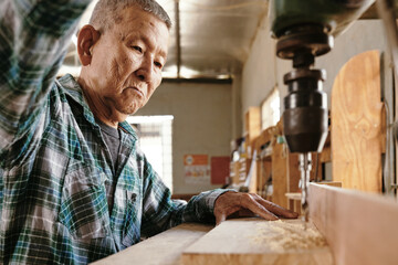 Elderly Asian carpenter using drilling machine to cut clean holes in wooden board