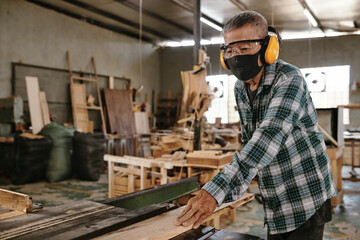Senior man in protective mask and earmuffs working with wood in carpenters workshop