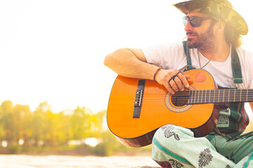 Hippy man with hat beard and glasses playing guitar outdoors on the beach