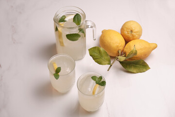 Pitcher and two of ice cold lemonade and lemons on the table. Refreshing mint lemonade in glasses on a white marble table. Top view.