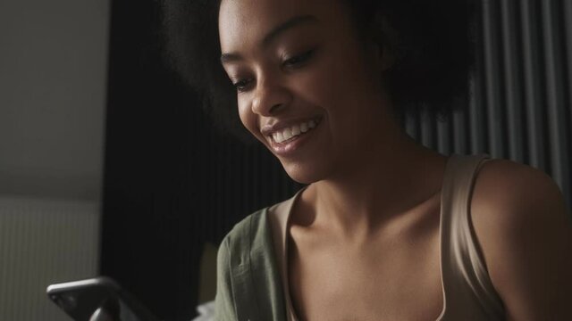 A black smiling woman looking at the phone while sitting on the bed in the room