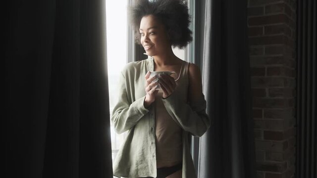 A pensive woman with a cup in her hands standing by the window in the room
