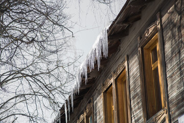 Icicles hang from the roof of a wooden house and shine in the sun. Against the background of a gray sky, branches of a tall tree. Abnormal weather concept. New Year's and Christmas is over.